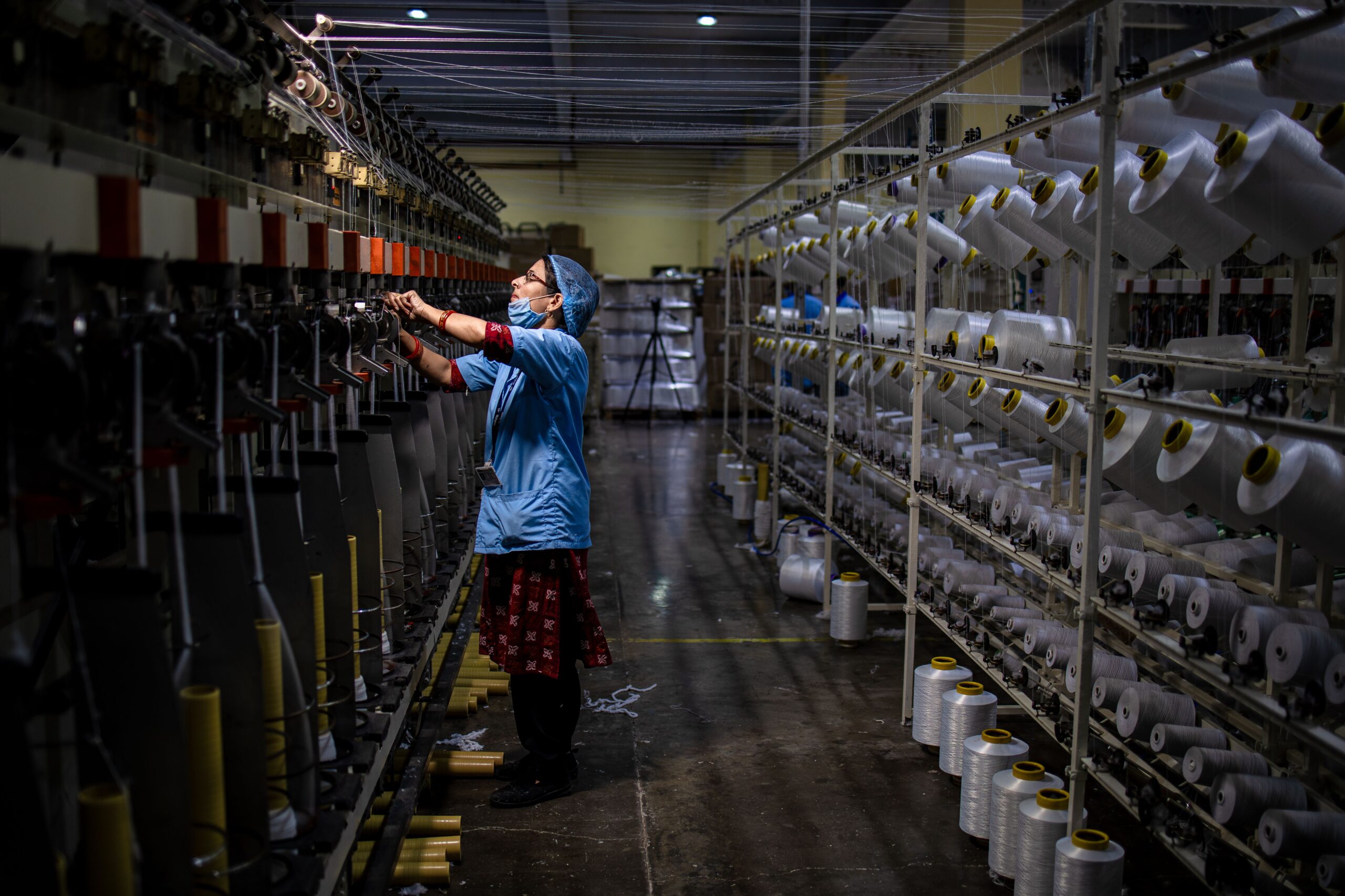 Masked female worker operating textile machinery in a factory setting. Indoor shot.